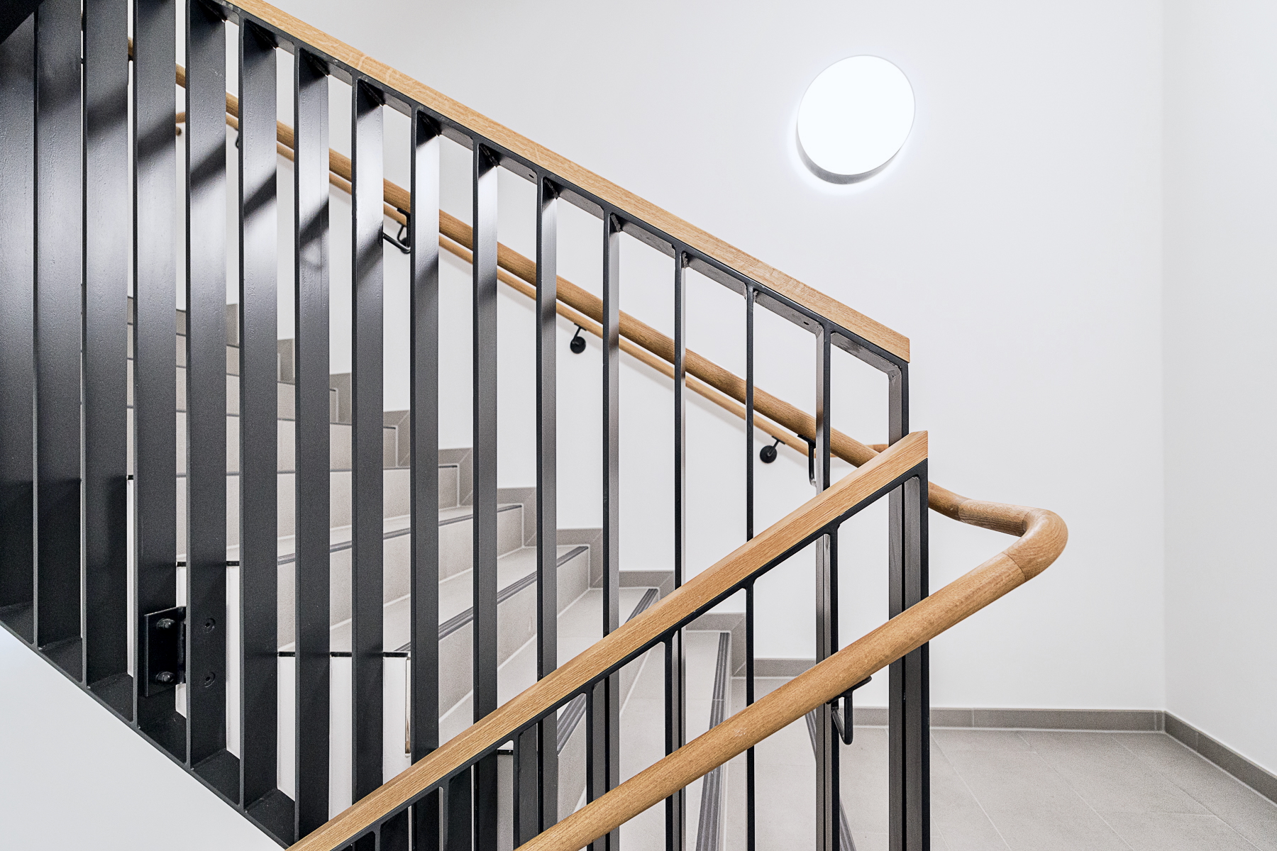 Detail des Treppengeländers im Finanzamt Waldmünchen, Außenstelle des Finanzamts Erlangen, mit Eichehandlauf, Eichenabdeckung des Metallstabgeländers, kontrastierenden Stufenvorderkanten und taktilen Schildern an den Handläufen.