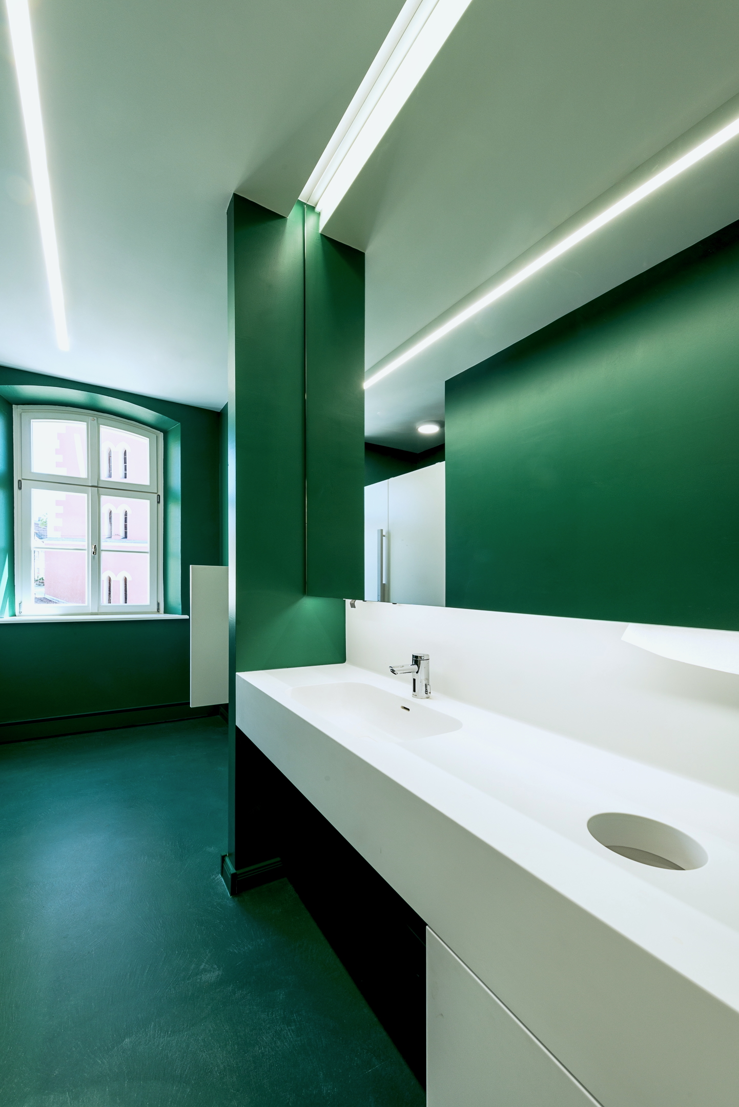 Sanitärbereich im Alten Amtsgericht Furth im Wald mit dunkelgrünem Boden und Wandflächen, Corianwaschtisch, indirekt beleuchtetem Spiegel und verdeckten Spendern.