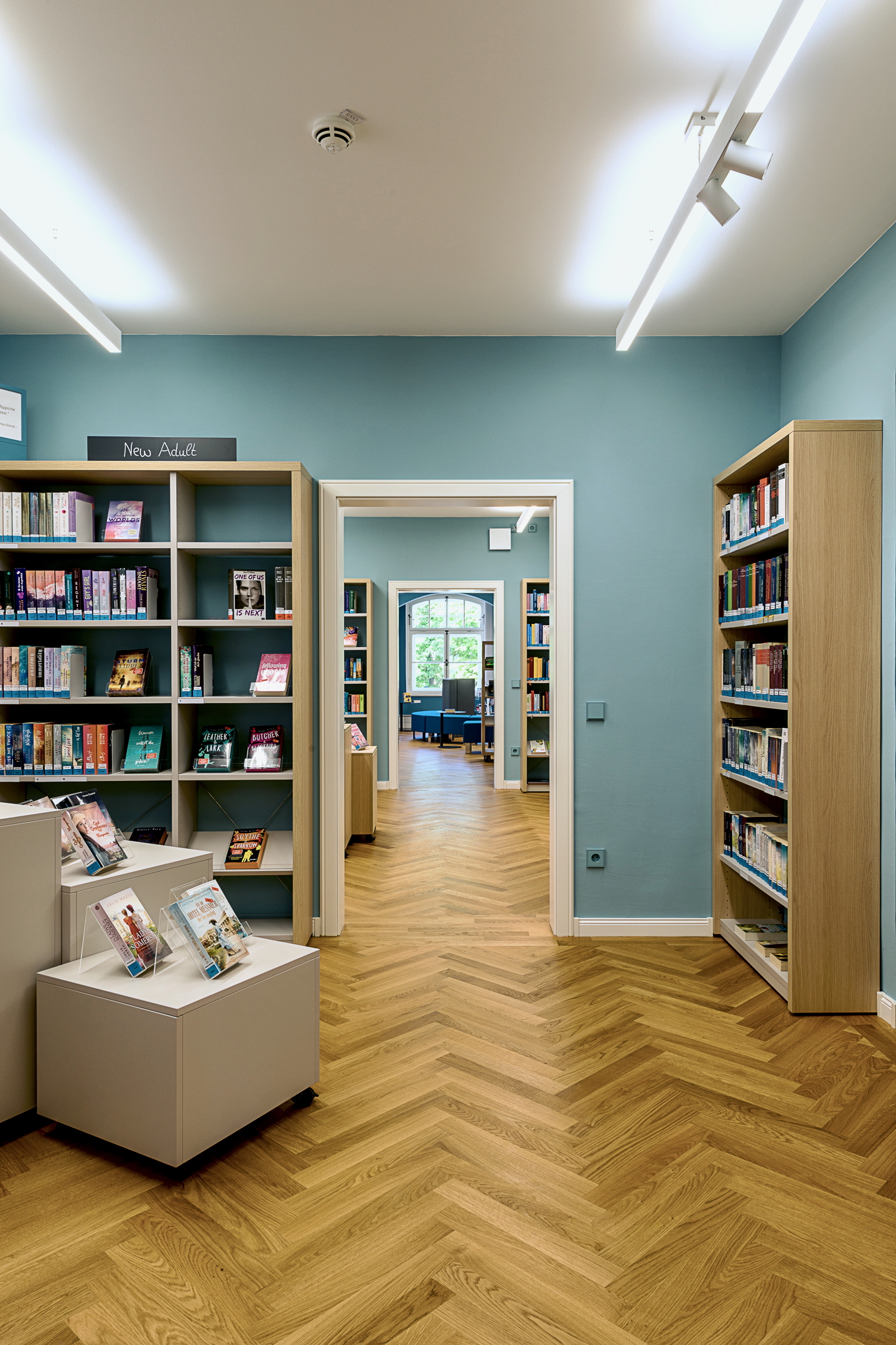 Bibliothek im Alten Amtsgericht Furth im Wald, Erwachsenenbereich mit Blick durch mehrere hintereinander angeordnete Türöffnungen, blaue Wandflächen, Eiche-Parkett, abgestimmte Bibliotheksmöblierung und farblich angepasstes Schalterprogramm.