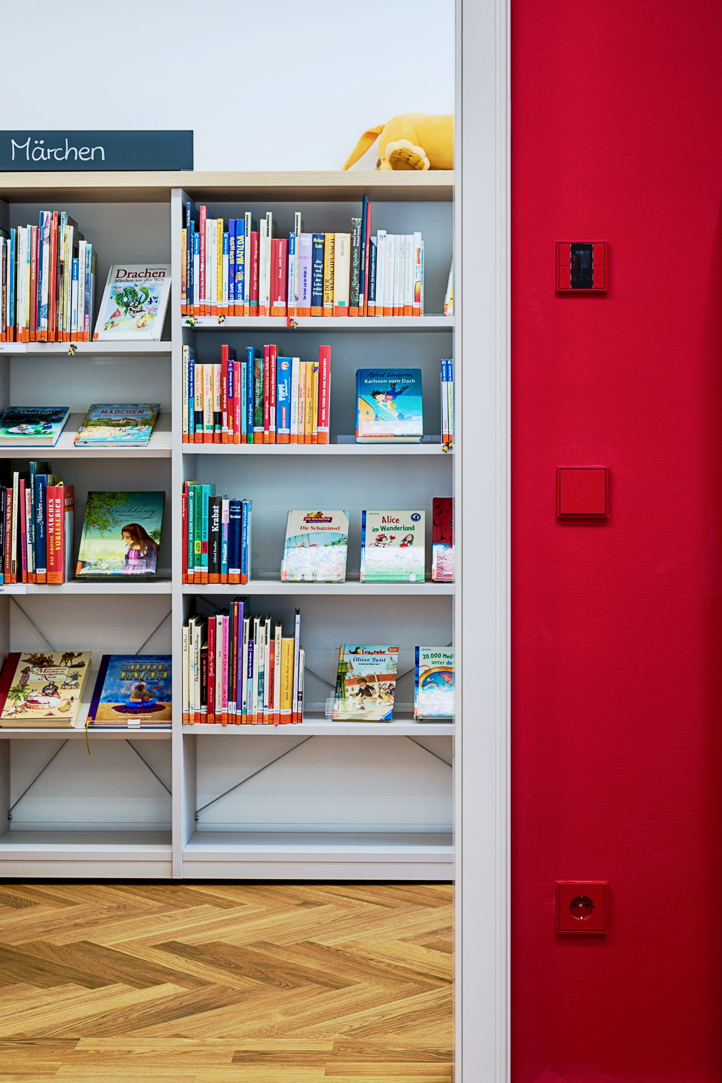 Detail einer Türleibung im Alten Amtsgericht Furth im Wald, weiße Leibung mit angrenzender Wand in kräftigem Rot, farblich abgestimmtem Schalterprogramm, Eiche-Parkettboden und im Hintergrund Bibliotheksmöblierung mit Kinderbüchern.