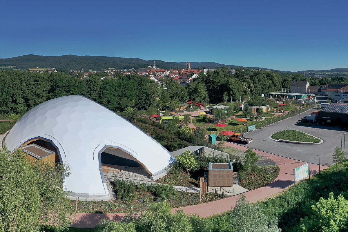 Drohnenaufnahme der Park-Arena Furth im Wald von Südosten, eingebettet in das Gelände der Landesgartenschau 2025, mit Blick auf Stadtturm, Kirche im Ortskern und den Voithenberg als Herkunftsort des Bauholzes, rechts im Bild die Drachenhöhle.