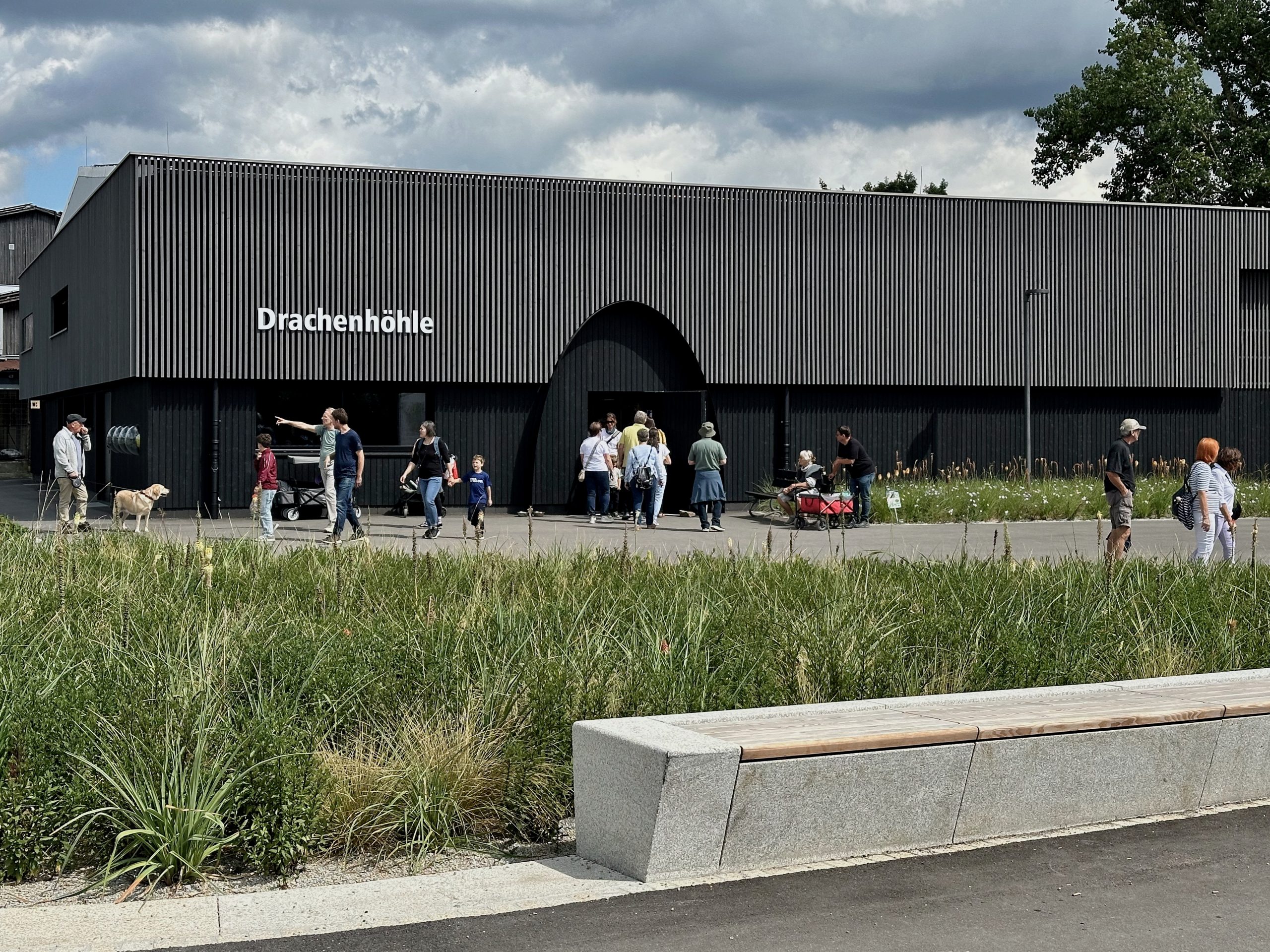 Südansicht der Drachenhöhle Furth im Wald mit verstecktem Eingang, carbonisierter Holzfassade im Sockelbereich und vorvergrauter Holzlattung oben.