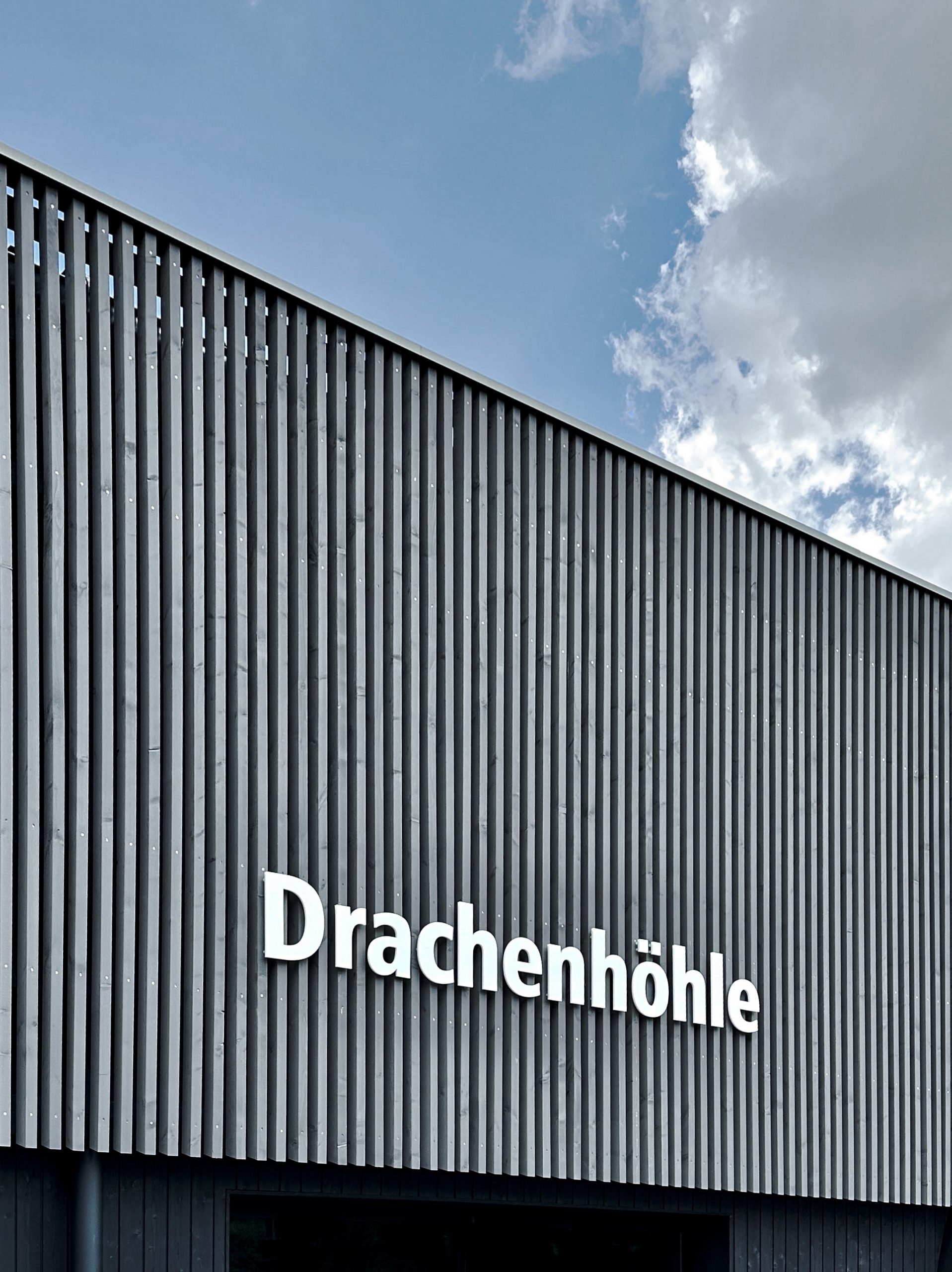 Blick nach oben auf die Fassade der Drachenhöhle Furth im Wald mit vorvergrauter Holzlattung und Schriftzug „Drachenhöhle“.