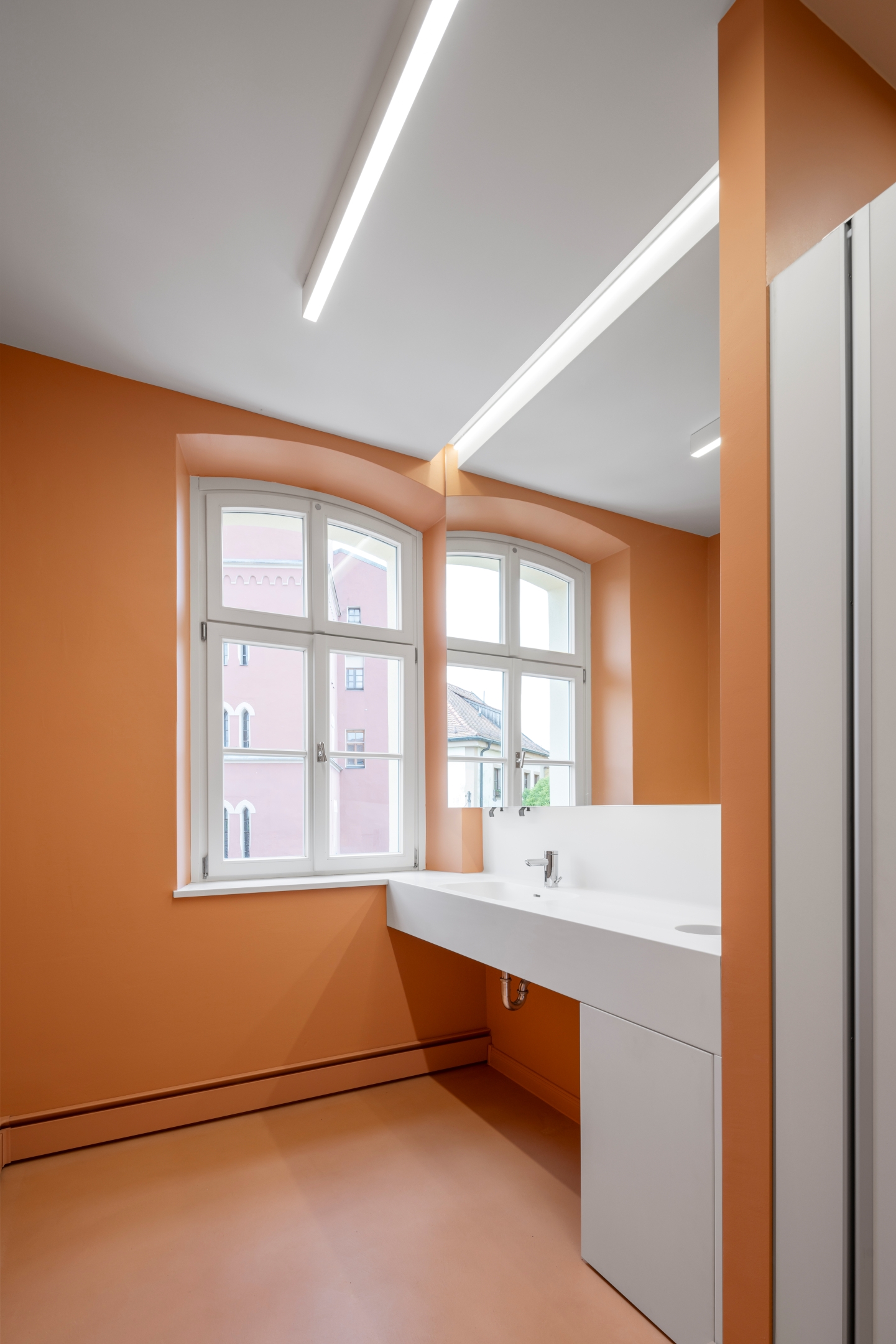 Sanitärbereich im Alten Amtsgericht Furth im Wald mit Apricot-Nude Boden und Wandflächen, Corianwaschtisch, indirekt beleuchtetem Spiegel und verdeckten Spendern.