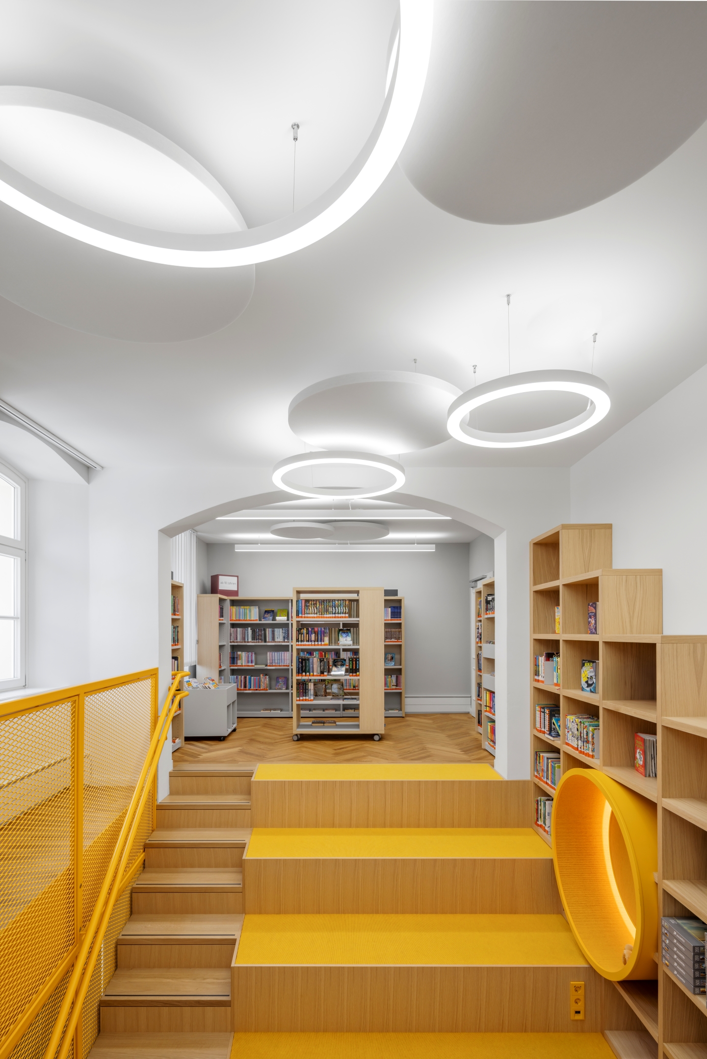 Sitztreppe aus Eiche im Alten Amtsgericht Furth im Wald mit gelbem Teppichbelag auf den Sitzflächen, integriertem Wandregal mit beleuchteten Sitzkreisen, gelbem Metallgeländer, farblich abgestimmtem Schalterprogramm und zusätzlicher Verbindung zwischen Erdgeschoss und Obergeschoss.