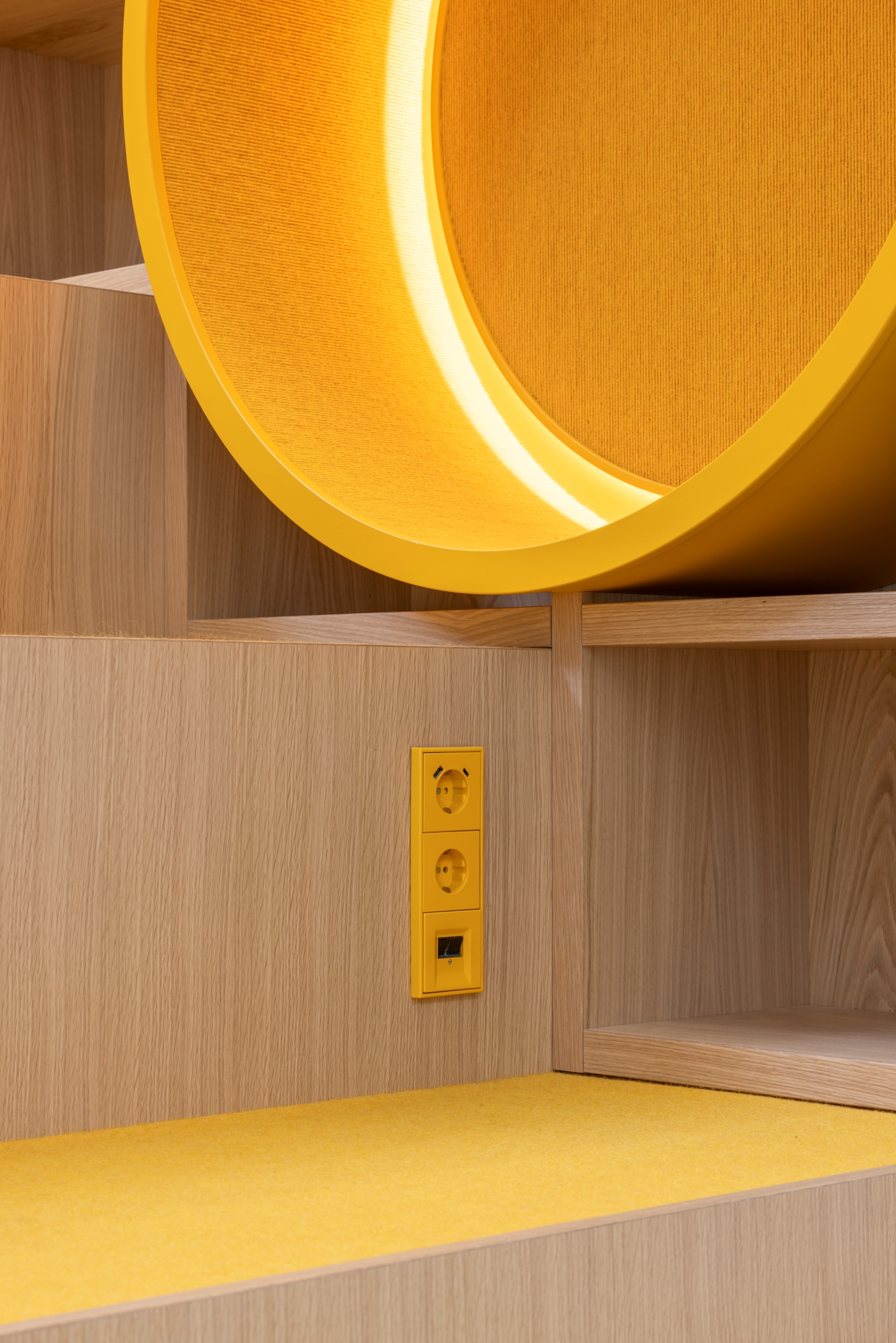 Detail der Sitztreppe im Alten Amtsgericht Furth im Wald mit Sitzfläche aus Eiche, gelbem Teppichbelag, integriertem Sitzkreis für Kinder und farblich abgestimmtem Schalterprogramm.