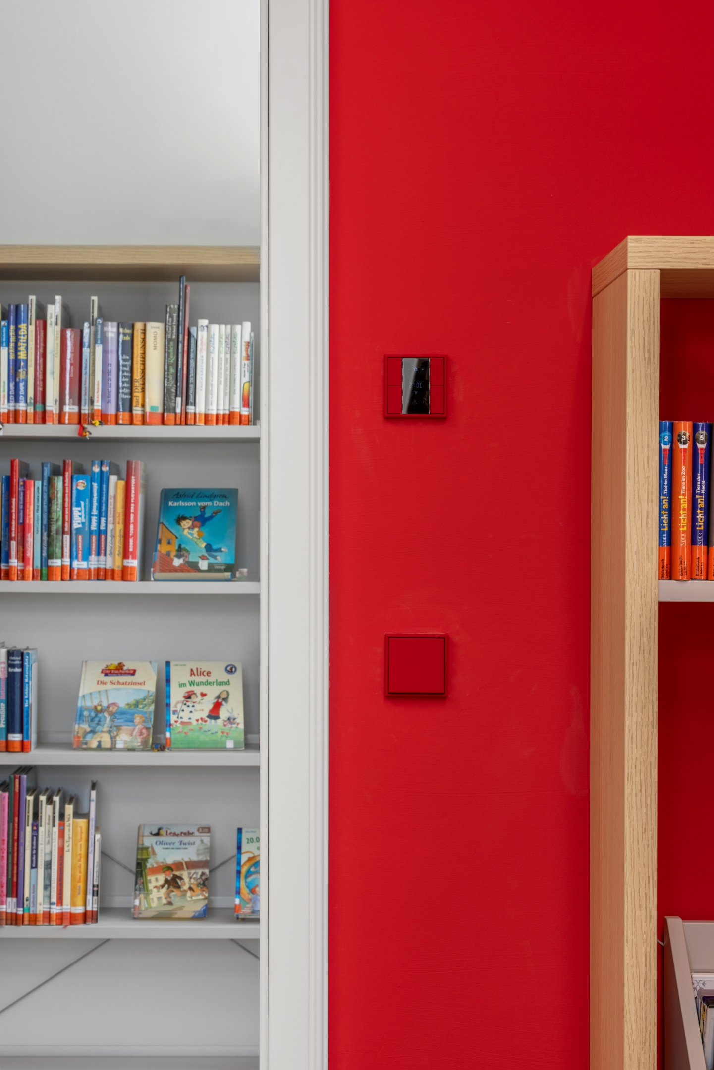 Detail einer Türleibung im Alten Amtsgericht Furth im Wald, weiße Leibung mit angrenzender Wand in kräftigem Rot, farblich abgestimmtem Schalterprogramm, Eiche-Parkettboden und im Hintergrund Bibliotheksmöblierung mit Kinderbüchern.